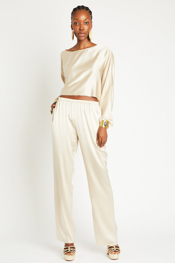 A model wearing a matching set of silk pants in a latte color with a relaxed fit and straight-leg style, paired with a silk top, standing against a white background.